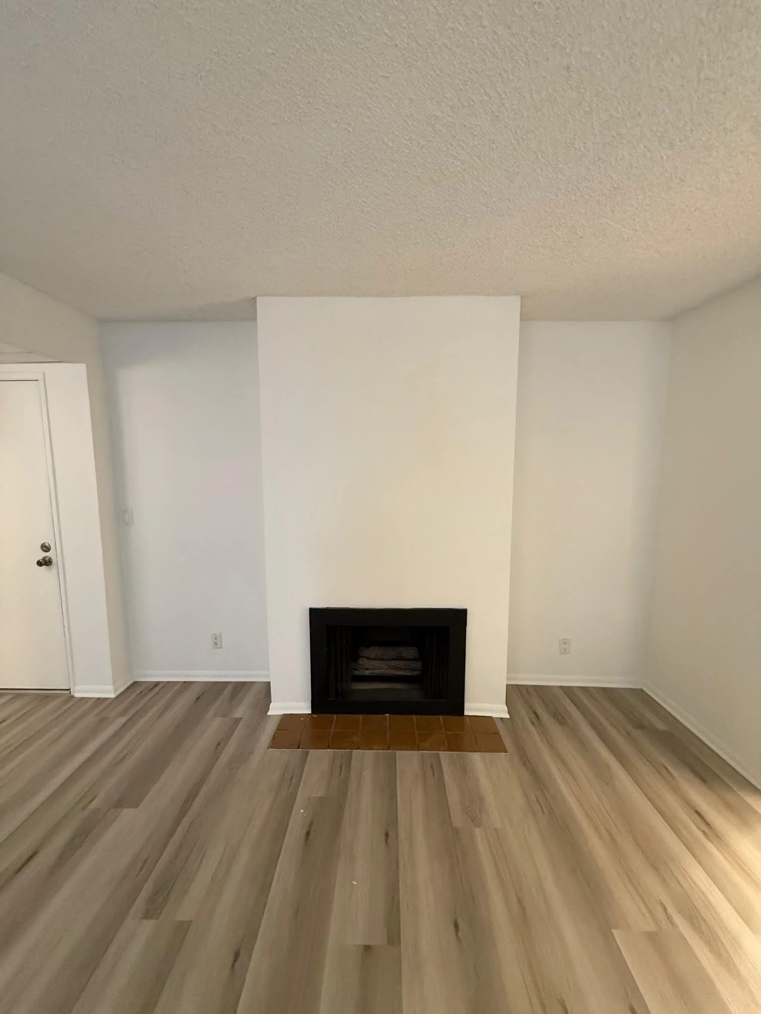 Living room with light wood flooring, white walls, a centered fireplace with a black opening, and a white column to the right of the fireplace.