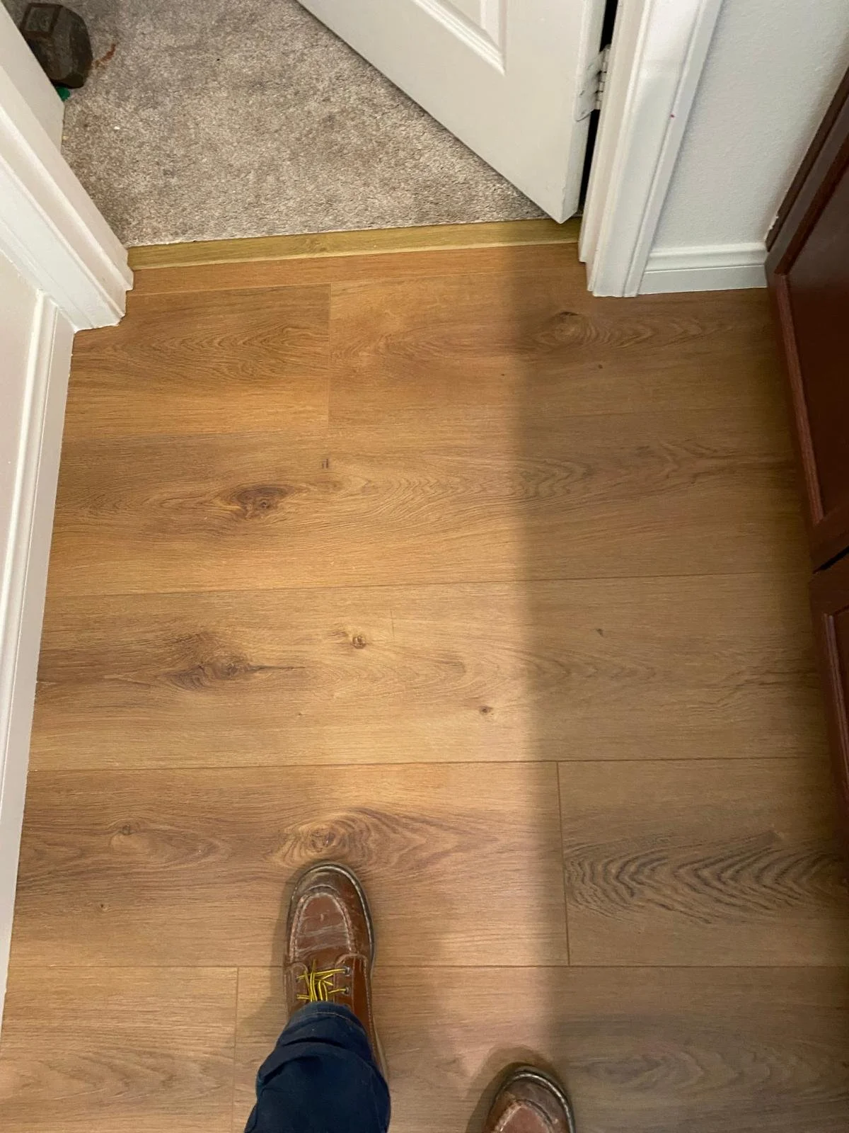 A person's feet in brown boat shoes and blue jeans are standing on a medium-toned hardwood floor. The photo is taken from the person's perspective, looking down at their feet. Part of a doorway with a threshold is visible, leading to a carpeted room.