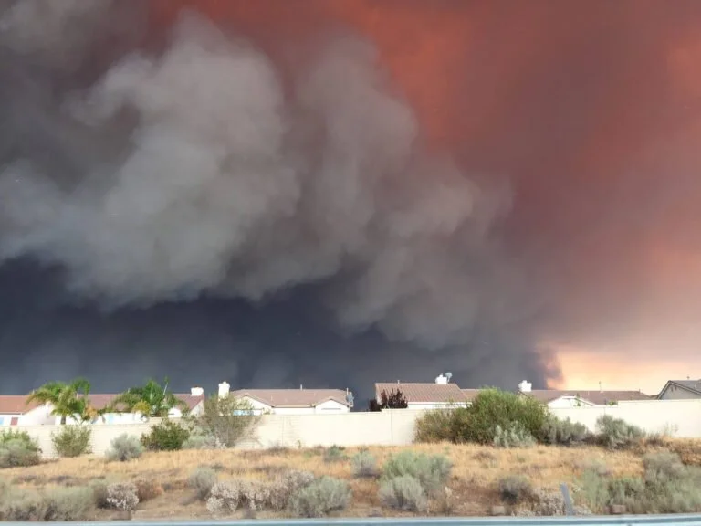Large dark smoke cloud in the sky over residential area with houses and desert vegetation, possibly from a fire.