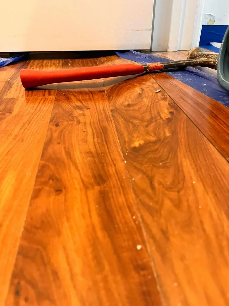 A hammer lying on a wooden floor near a wall and door, with a blue painter's tape on the floor.