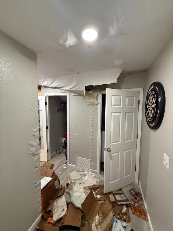 Ceiling collapsed in a hallway, exposing insulation and damage. Debris, boxes, and trash on the floor. Open closet doors and a dartboard on the wall.