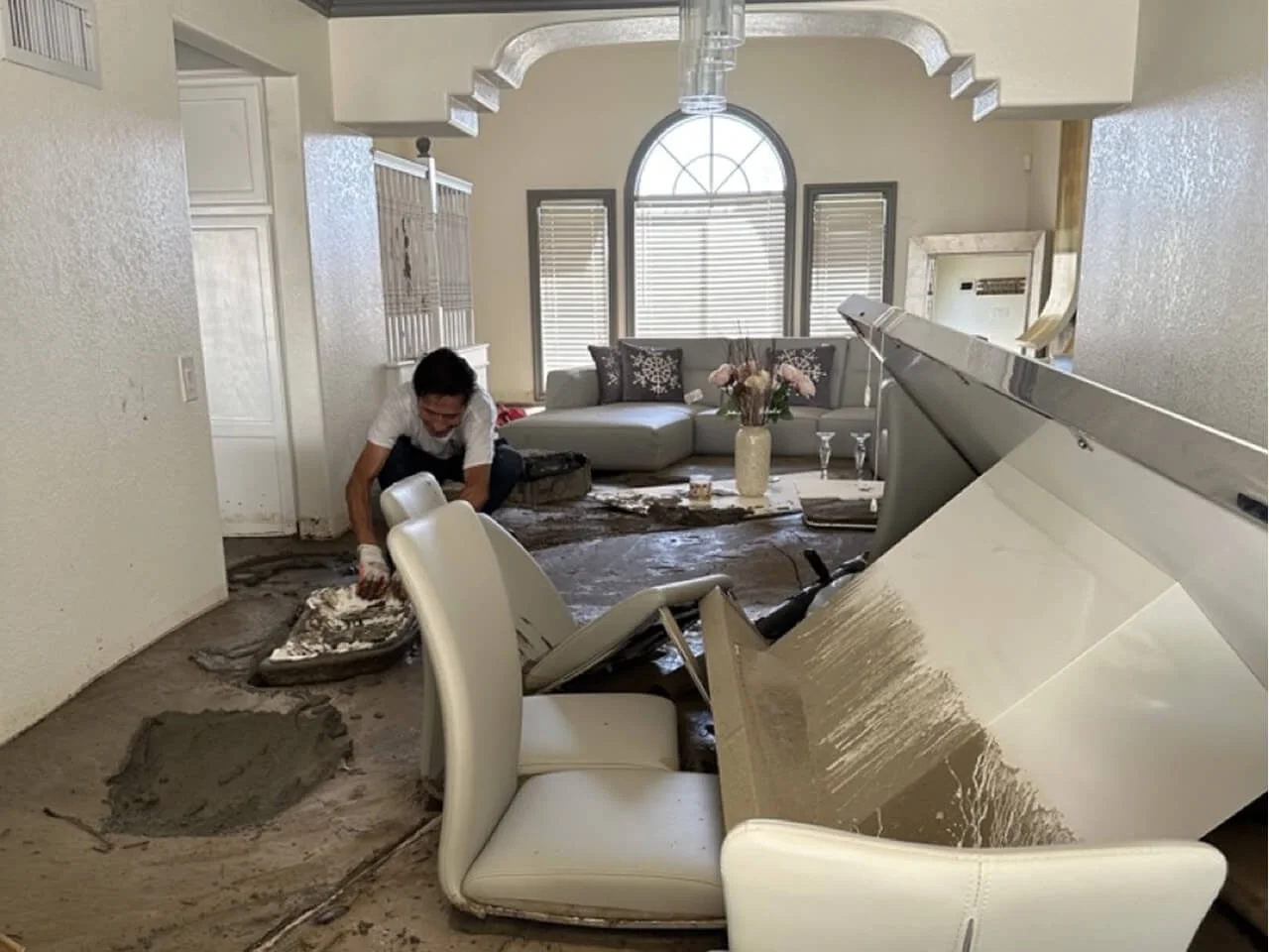 A person cleaning up a flooded living room with a white grand piano and a gray sofa. Water and mud are on the floor and furniture.