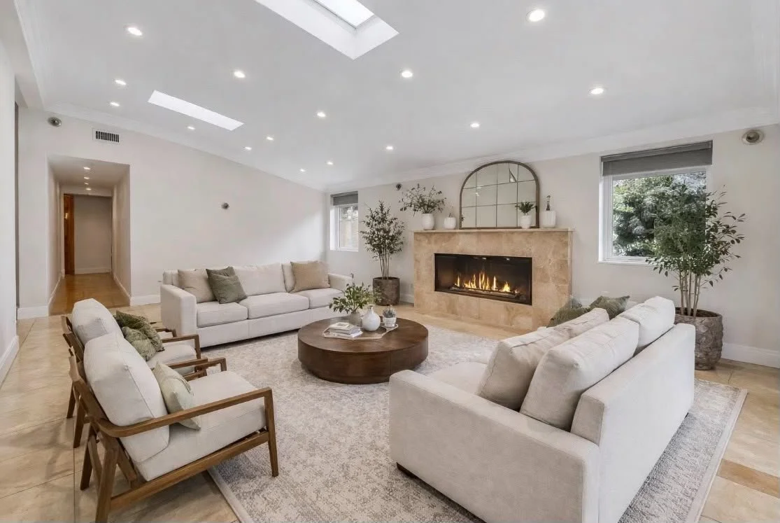 Modern living room with beige sofas, a round wooden coffee table, a fireplace with a beige stone surround, and large potted plants, illuminated by recessed ceiling lights.