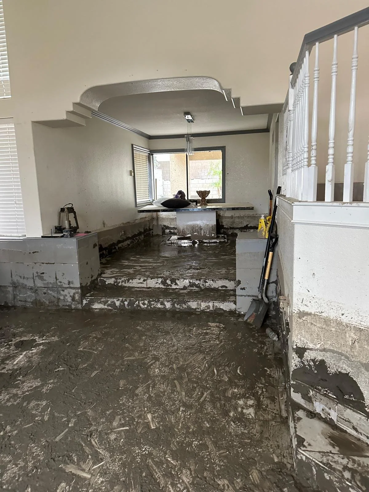 Interior of a house under renovation with part of the floor and steps covered in wet concrete, tools and construction materials scattered around, and a window at the back with sunlight coming through.
