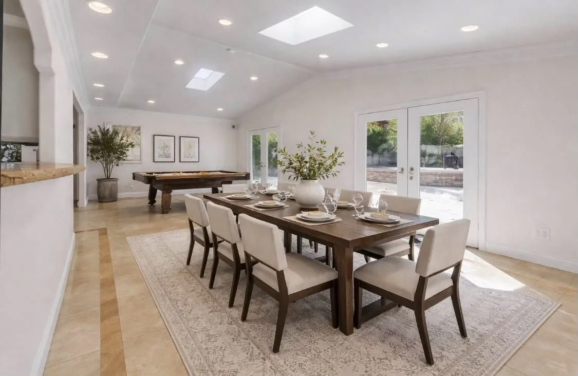 Dining room with a large wooden table set for six, featuring white chairs, a white vase with green foliage, and a beige rug, with a pool table and framed artwork on the white walls in the background.
