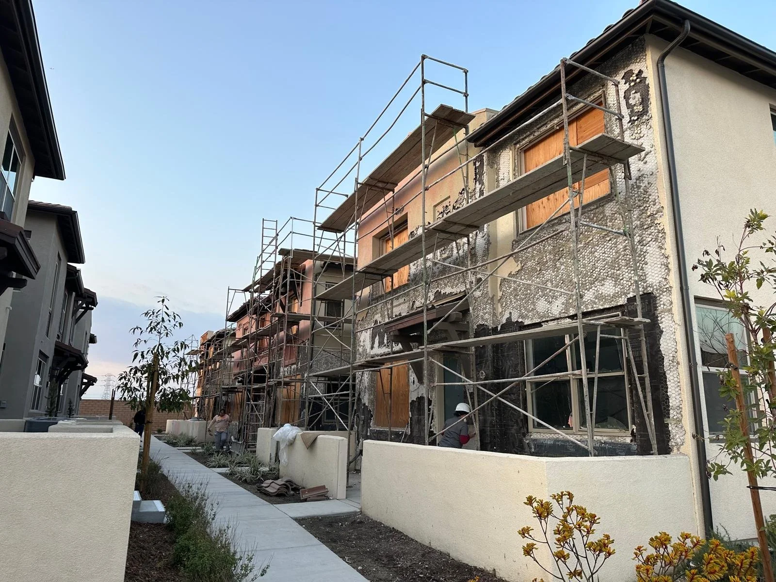 Construction workers building or renovating a house with scaffolding surrounding the structure, some windows boarded up, and a partly damaged exterior wall.