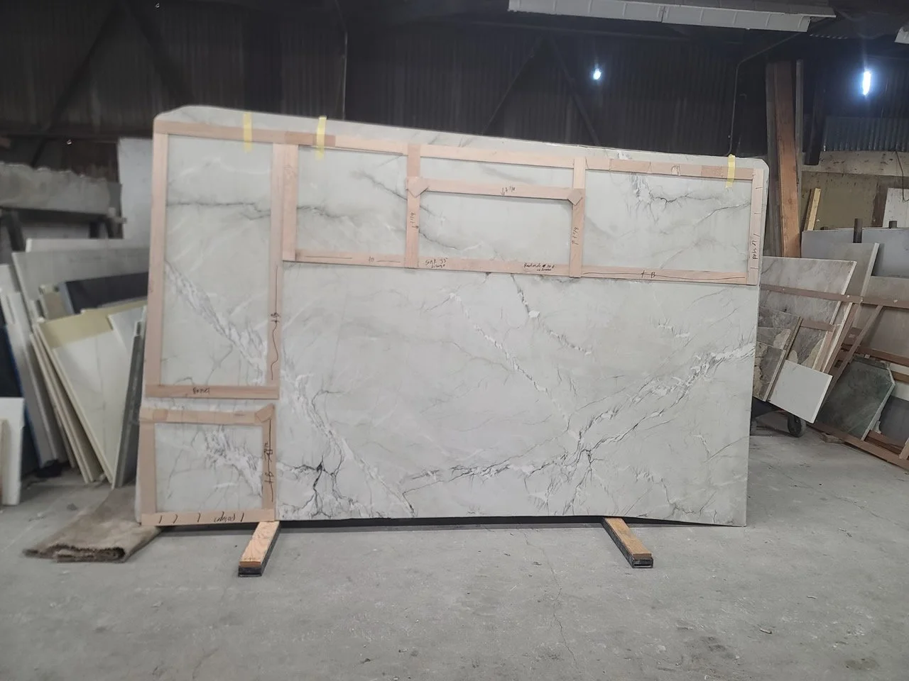 Large marble slab with wooden framing and measurements, standing in a warehouse with other stone slabs in the background.