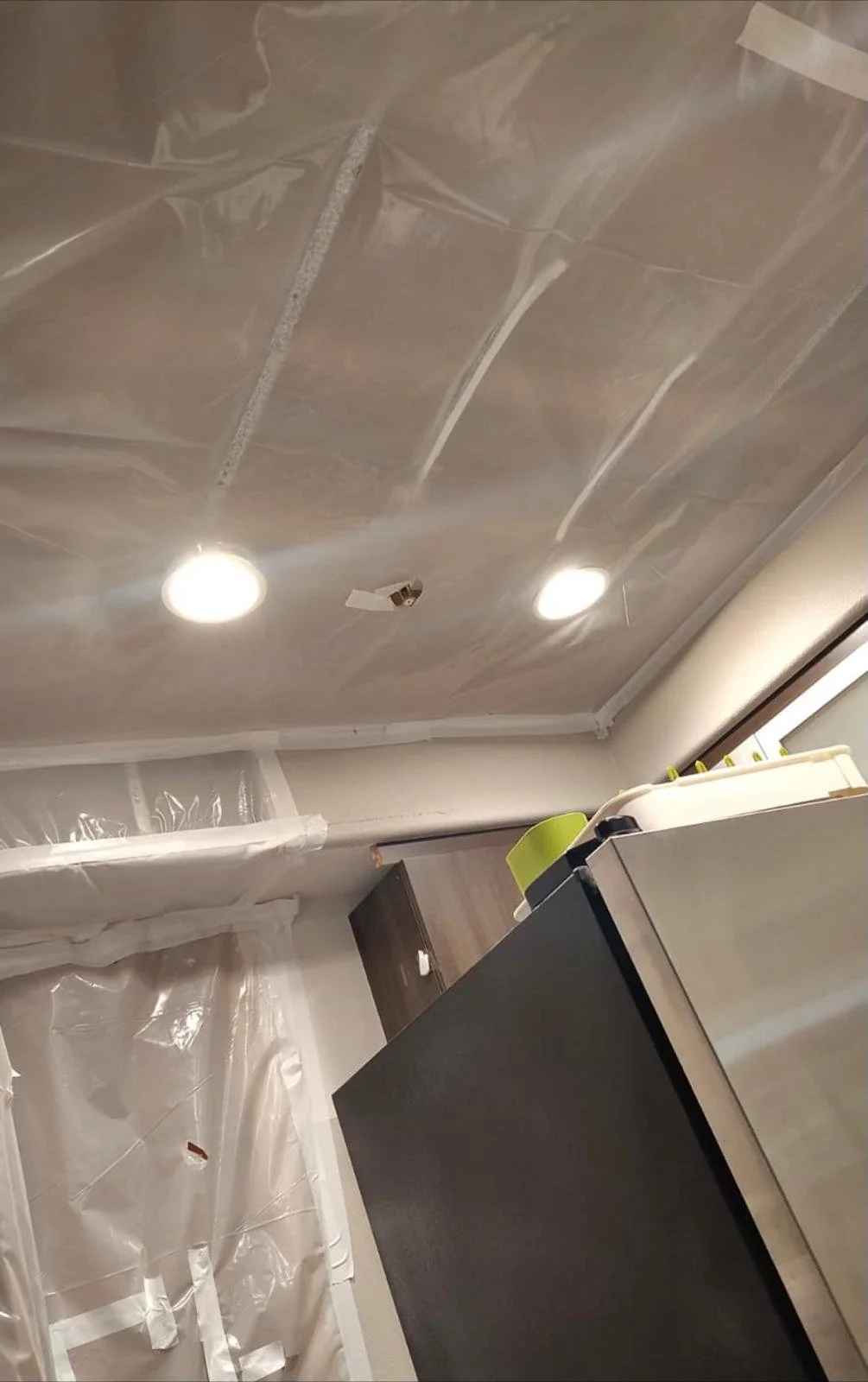 Ceiling with recessed lighting, covered with plastic sheeting for painting or renovation, part of a kitchen with a black refrigerator and small upper cabinets.