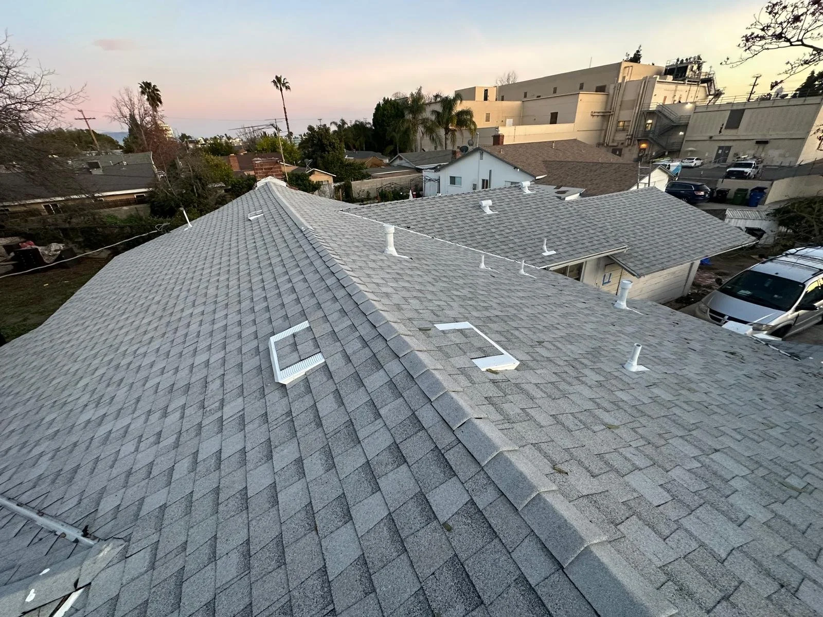 A view of residential rooftops with gray shingles, several roof vents, and skylights, with trees, neighboring houses, and a larger building in the background, during sunset.
