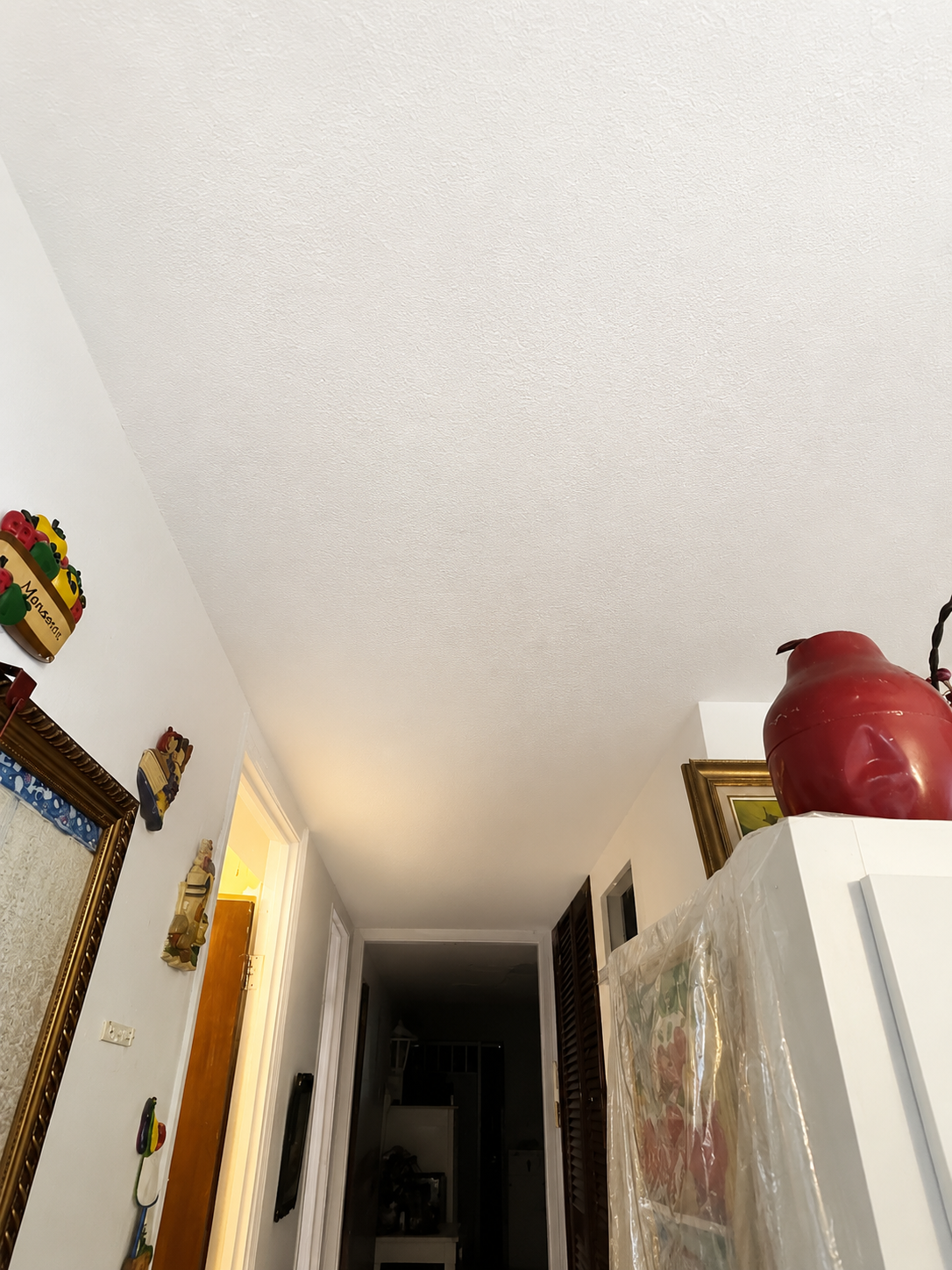 View of a narrow hallway in a home with white textured ceiling and walls decorated with small figurative artwork; partially visible doorway with illuminated room; a large red pitcher on top of a white covered appliance with framed artwork nearby.