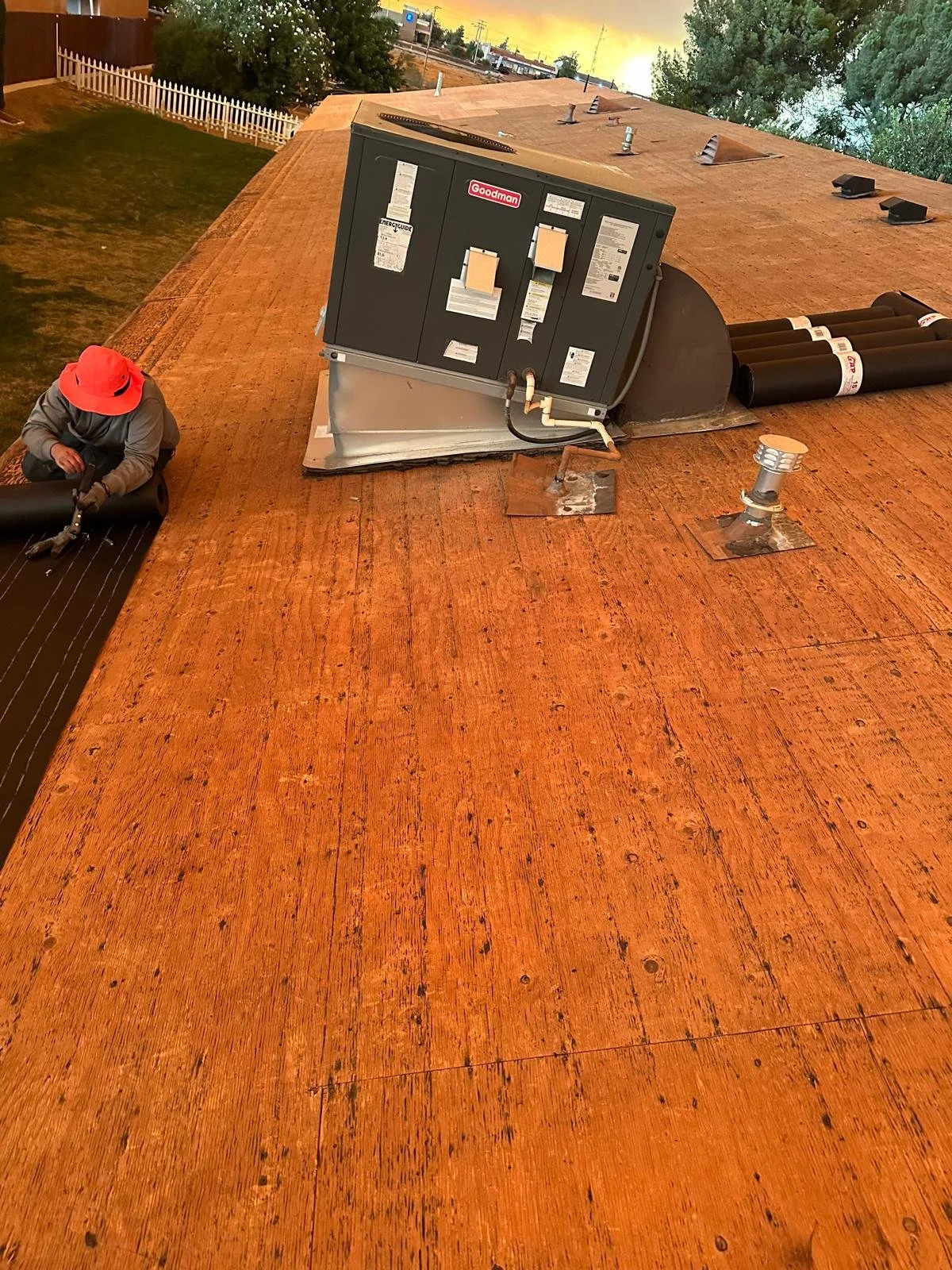 A roofing worker installing black underlayment on a flat, orange wooden roof. An industrial HVAC unit is mounted on the roof, with pipes and vents. The sky in the background is orange with trees and power lines visible.