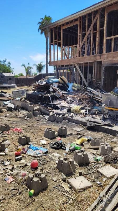 Debris and trash scattered in front of a damaged, partially built wooden house with exposed framework, under a clear blue sky with palm trees in the background.
