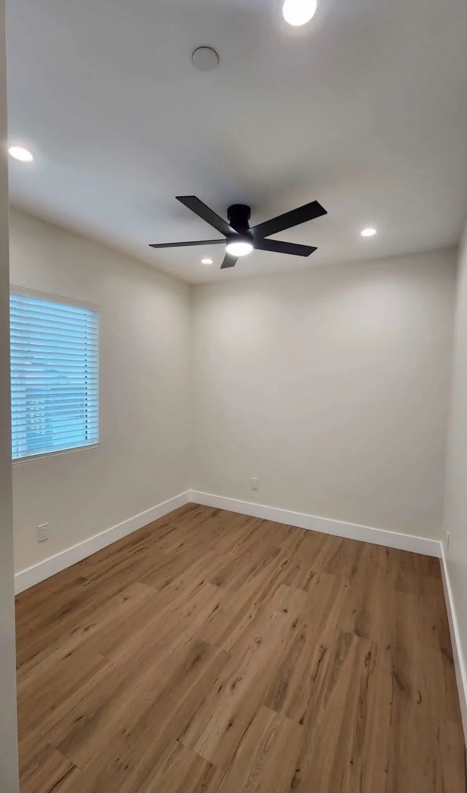 Empty room with beige walls, wooden floor, ceiling fan, ceiling lights, and a window with blinds.