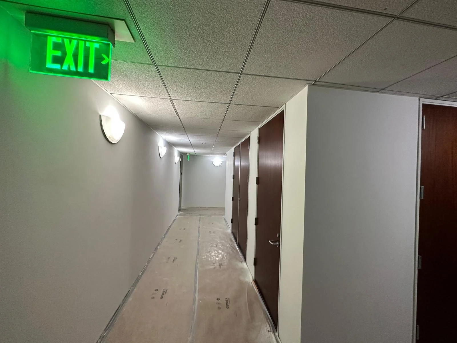A hallway with a green illuminated exit sign on the ceiling. The hallway has white walls, round wall-mounted lights, and closed wooden doors along one side. The floor is covered with protective plastic sheeting.
