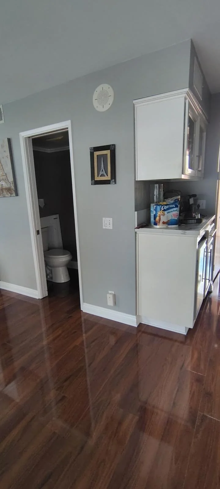 Corner of a kitchen with gray walls, white cabinets, a bathroom doorway with a toilet, and a wooden floor.