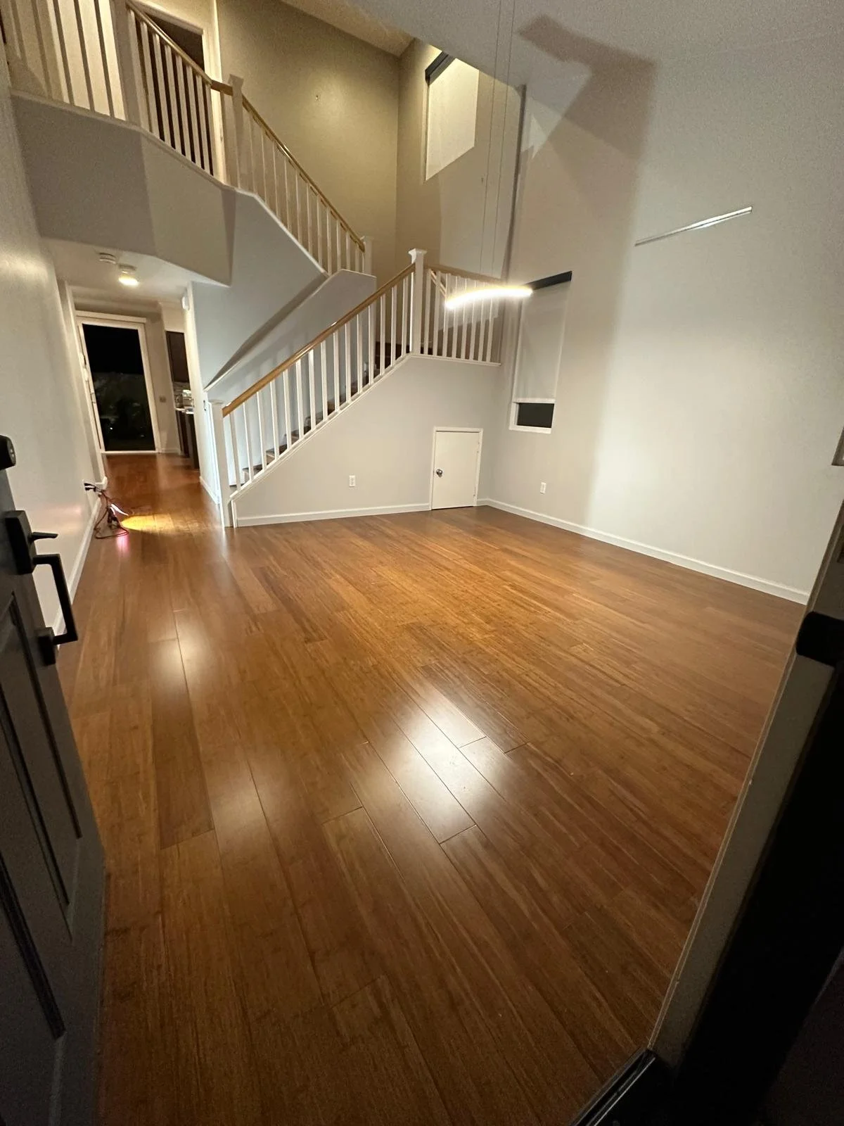 Interior of a living room with hardwood floors, a staircase with a white railing and wooden handrail, and a small door underneath the staircase.
