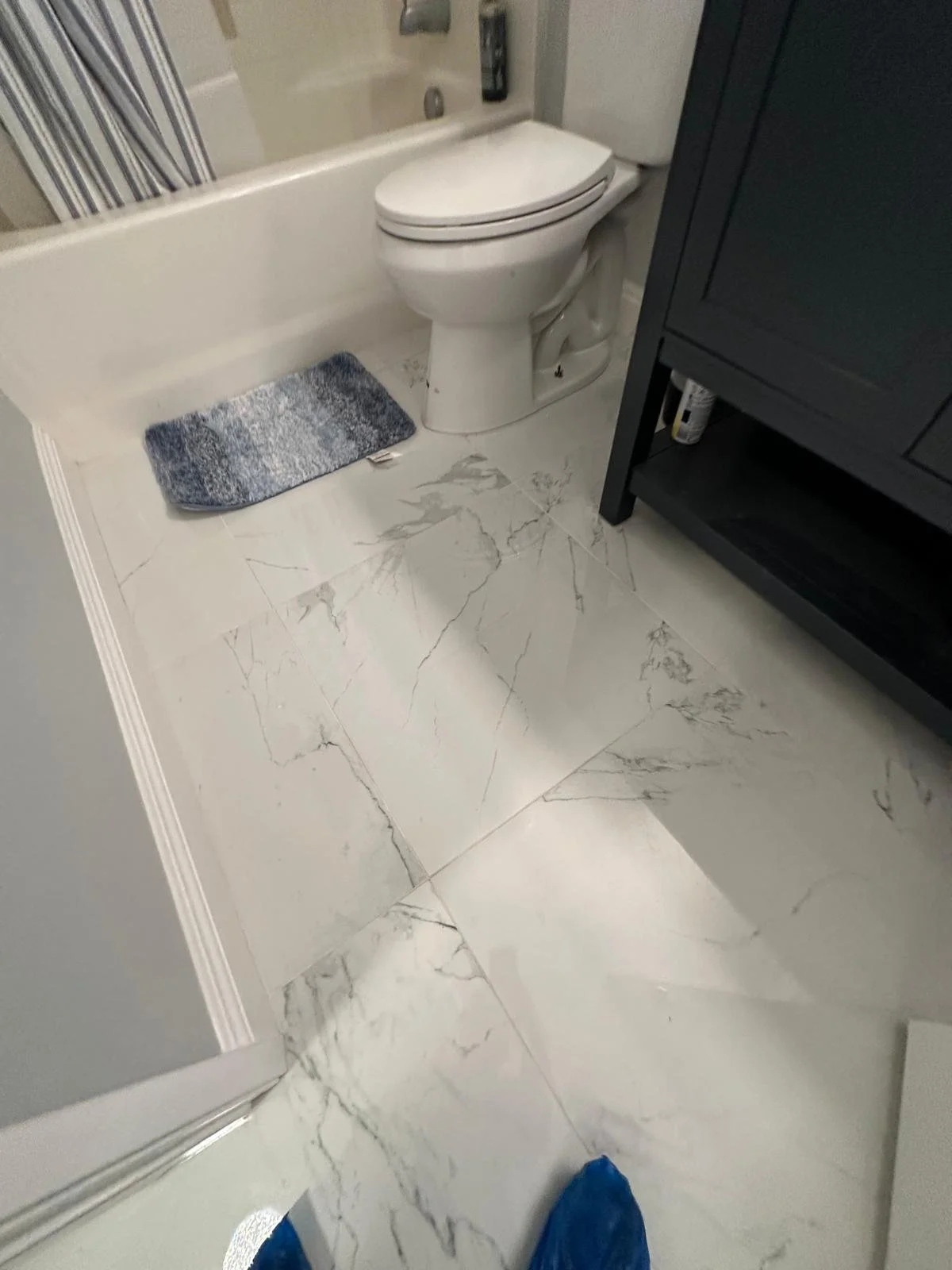 Photo of a bathroom with marble tile flooring, a white toilet, a small gray and white rug near the bathtub, and a black bathroom cabinet on the right side.
