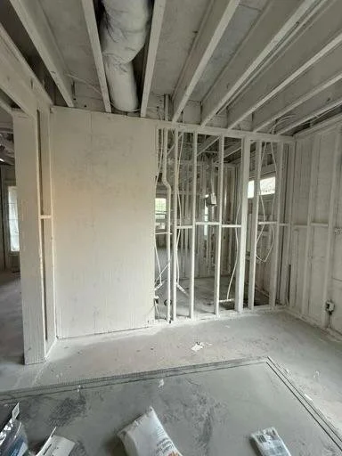 Interior of a building under construction with exposed drywall and framing, pipe visible on the ceiling, and construction materials on the floor.