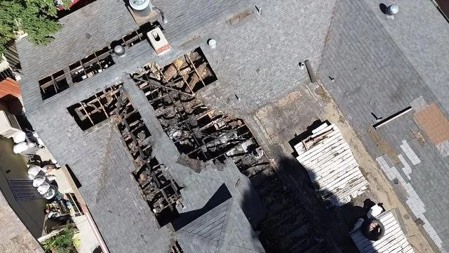 Aerial view of a roof with a large section that has been partially removed, exposing wooden beams underneath. The roof appears to be under repair, with some roofing materials and debris scattered around.