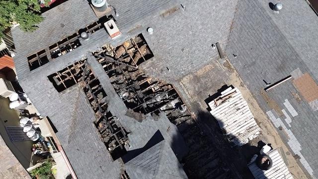 Aerial view of a roof with a large section that has been partially removed, exposing wooden beams underneath. The roof appears to be under repair, with some roofing materials and debris scattered around.