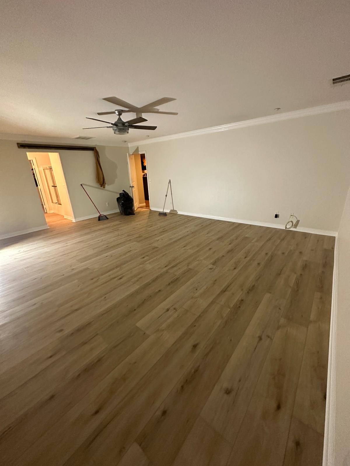 Empty living room with wooden flooring, ceiling fans, and cleaning tools near the wall.