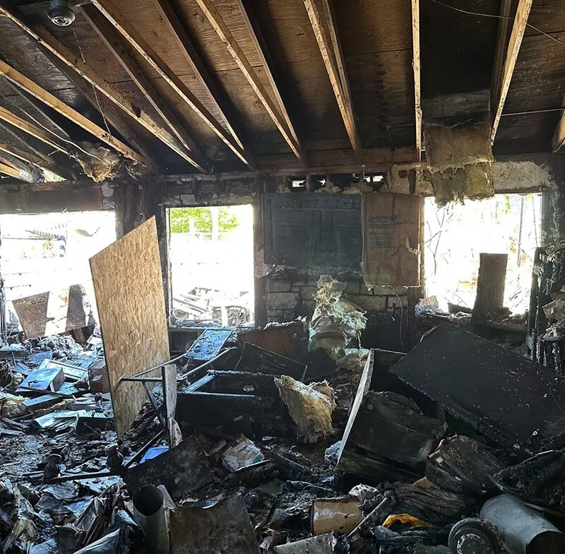 Interior of a severely damaged and smoke-filled room after a fire, with charred debris, broken furniture, and burned insulation, windows showing daylight outside.