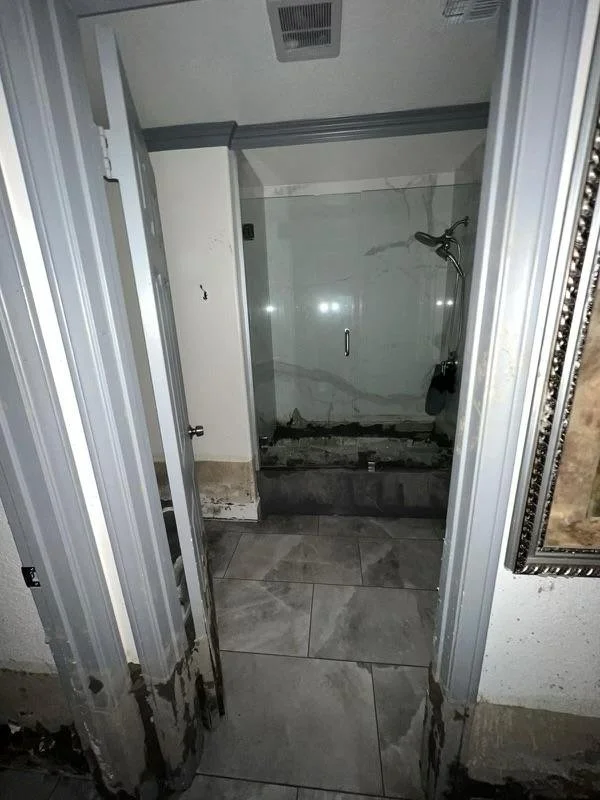 View of a damaged bathroom shower area with cracked tile wall, missing tiles near the floor, and a showerhead on the right side.
