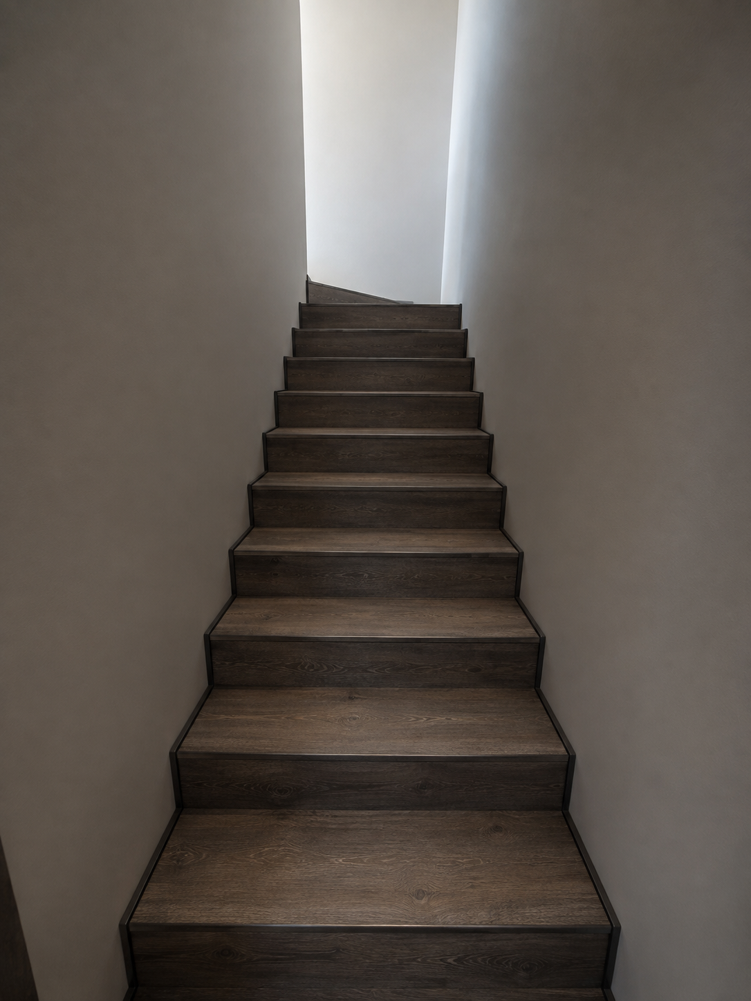 A narrow staircase with dark wooden steps leading upward, flanked by plain white walls, illuminated by natural light at the top.