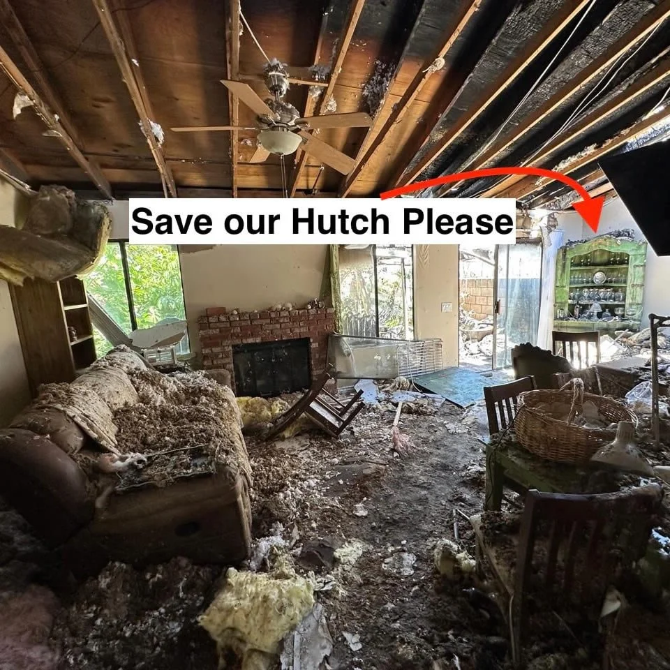 Living room with damaged ceiling, furniture covered in insulation, and debris scattered across the floor, indicating a recent fire or disaster.