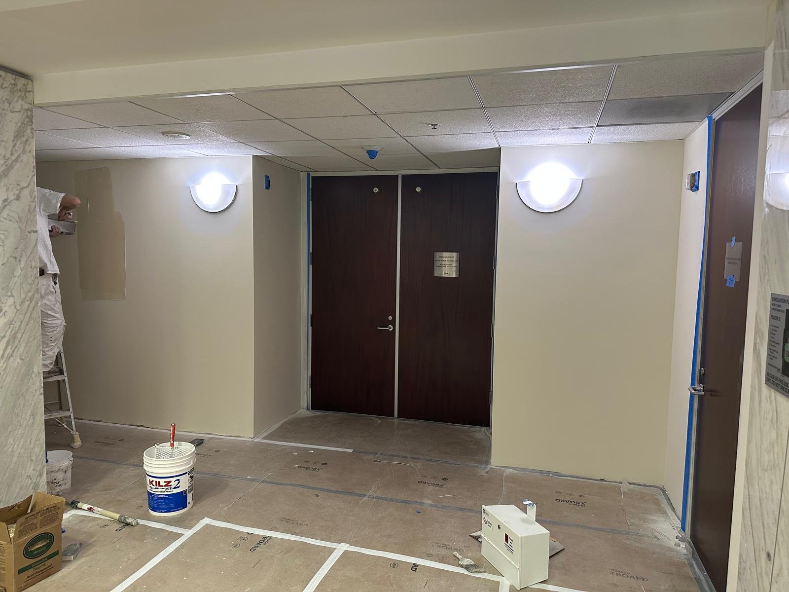 A hallway under construction with a closed dark wooden double door, two wall-mounted light fixtures, a person on a ladder, and construction materials on the floor.