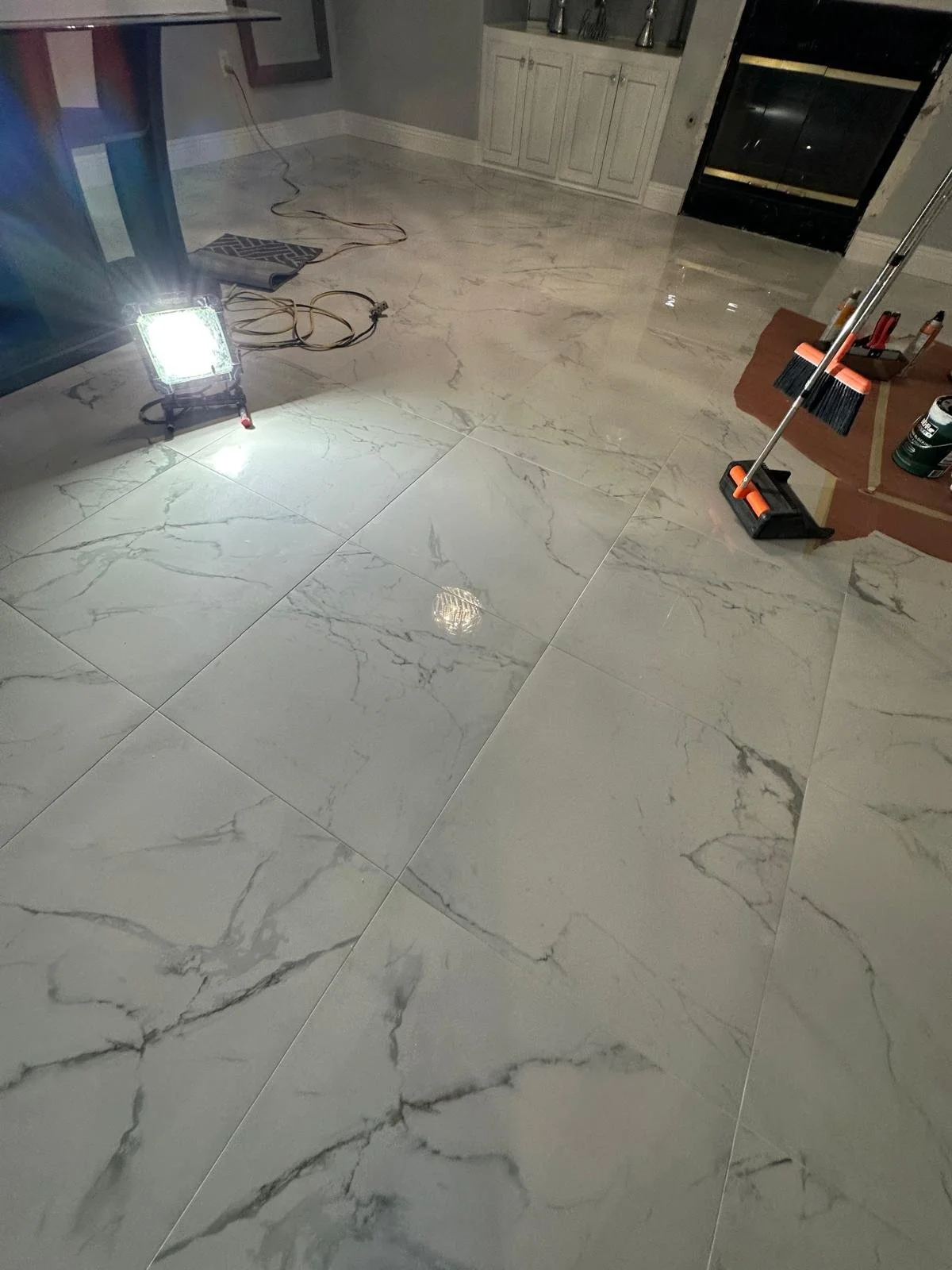 A room with large marble tiles on the floor, construction tools and lighting equipment present, and unopened cabinetry near a fireplace.