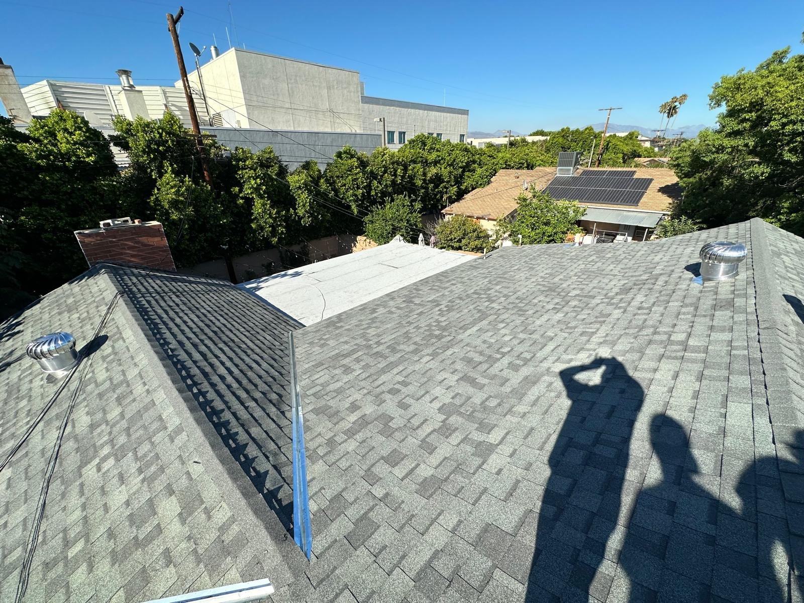 A view of rooftops with shingles, some with solar panels, in a neighborhood with trees and a large industrial building in the background. The photo was taken on a sunny day.