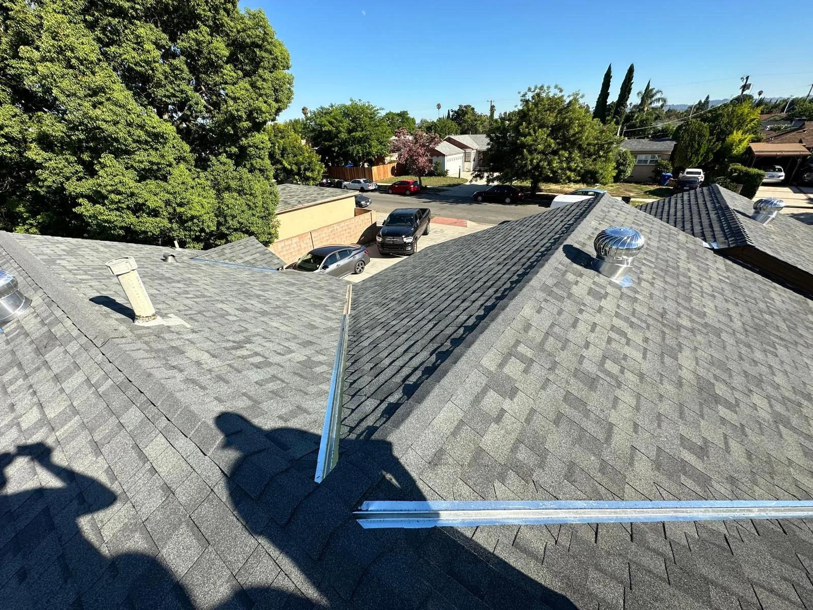 View from a rooftop showing gray shingle roof with metal vents and nearby trees, parked cars, and residential houses along the street.