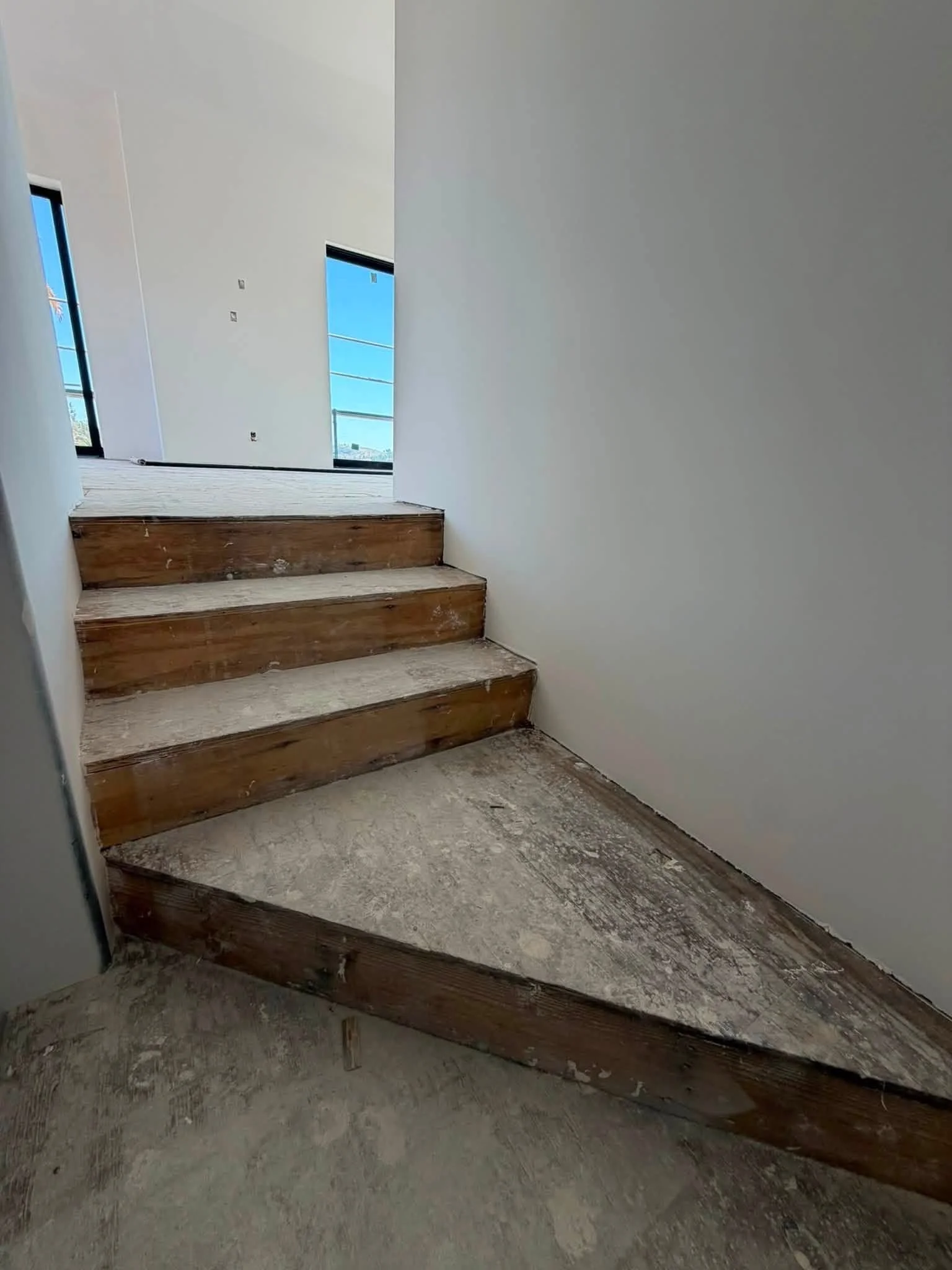 Interior of a house under construction with unfinished wooden stairs and white walls, no railing, and exposed concrete flooring.