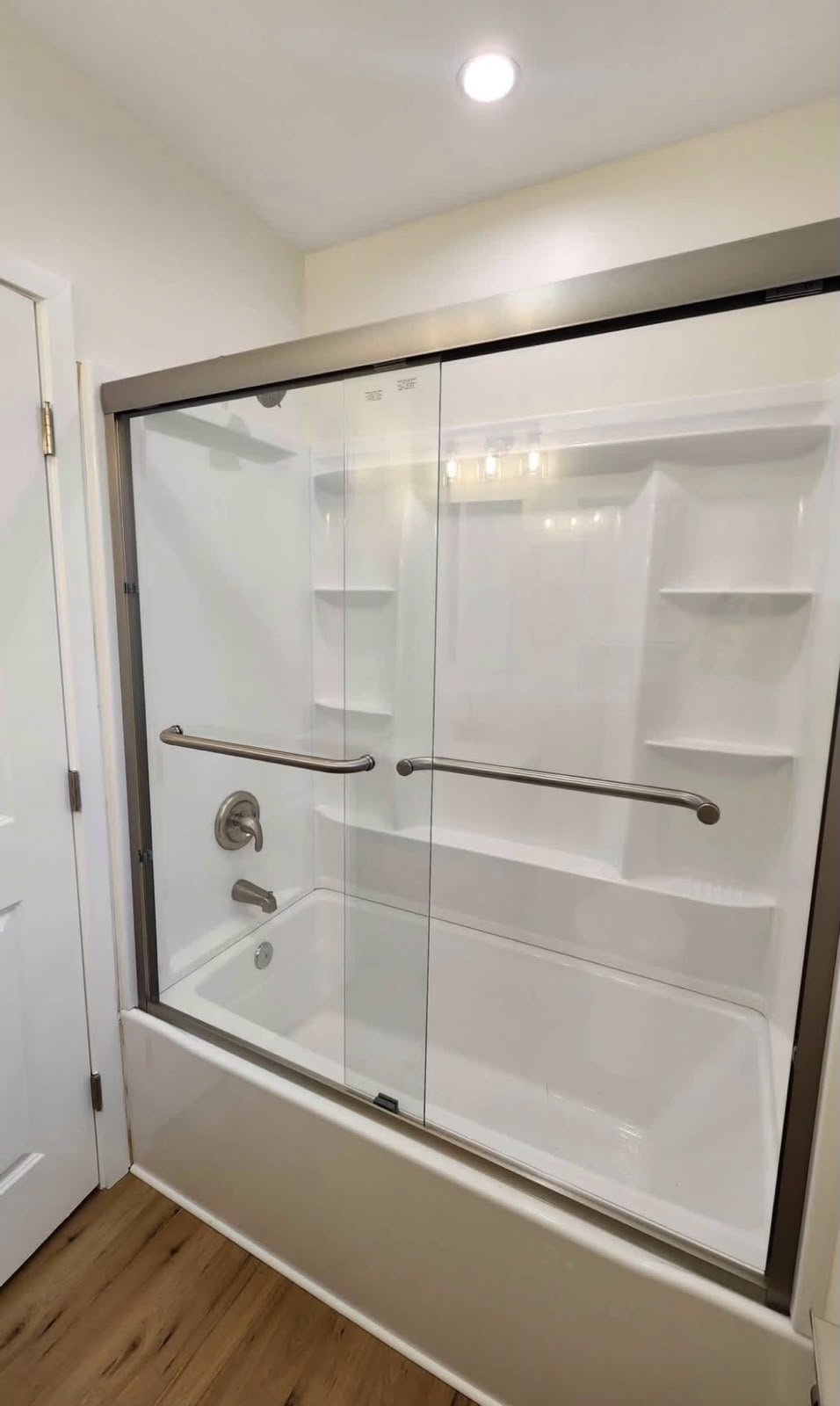 A walk-in shower with glass sliding doors, metal handles, and built-in shelves, situated in a bathroom with wooden flooring and white walls.