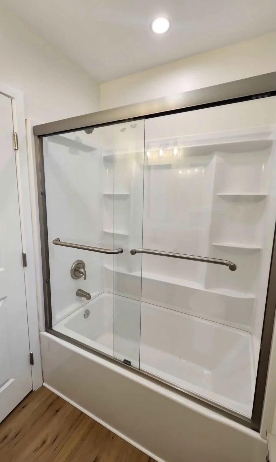 A walk-in shower with glass sliding doors, metal handles, and built-in shelves, situated in a bathroom with wooden flooring and white walls.