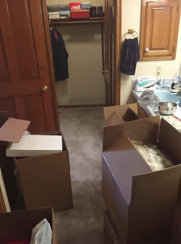 Kitchen with cardboard boxes on the floor, open closet with clothes hanging, and small kitchen appliances on the counter.