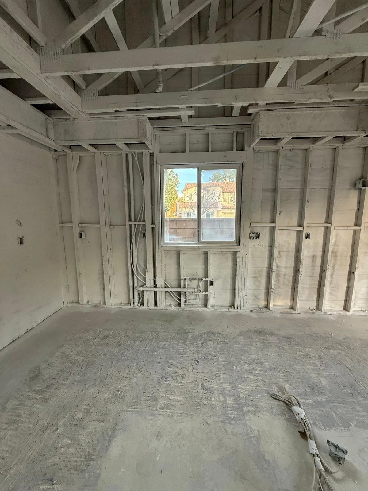 Unfinished room with drywall, wiring, and a window showing an outside view of houses and trees.