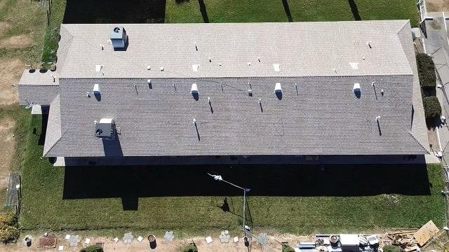 Aerial view of a single-story building with a light gray roof and multiple vents, surrounded by a green lawn and a paved patio with outdoor furniture and equipment.