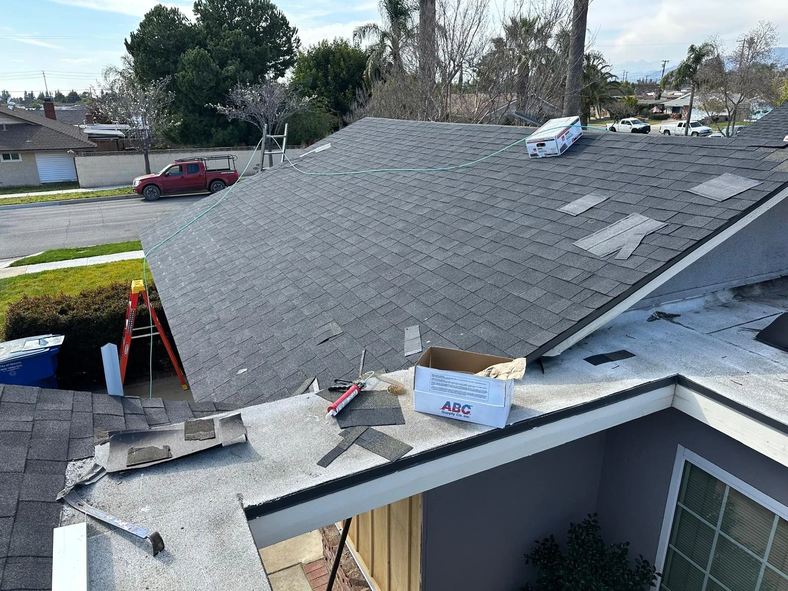 View of a house roof with shingles being repaired or replaced, tools, and materials on the roof, including a caulk gun, a box of supplies, and some shingles, with a ladder and nearby residential neighborhood in the background.