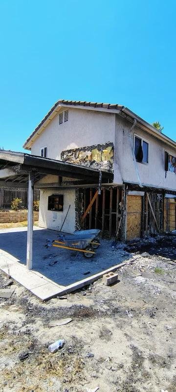 A two-story house with damage, missing siding, and an open basement area with construction materials outside under a clear blue sky.