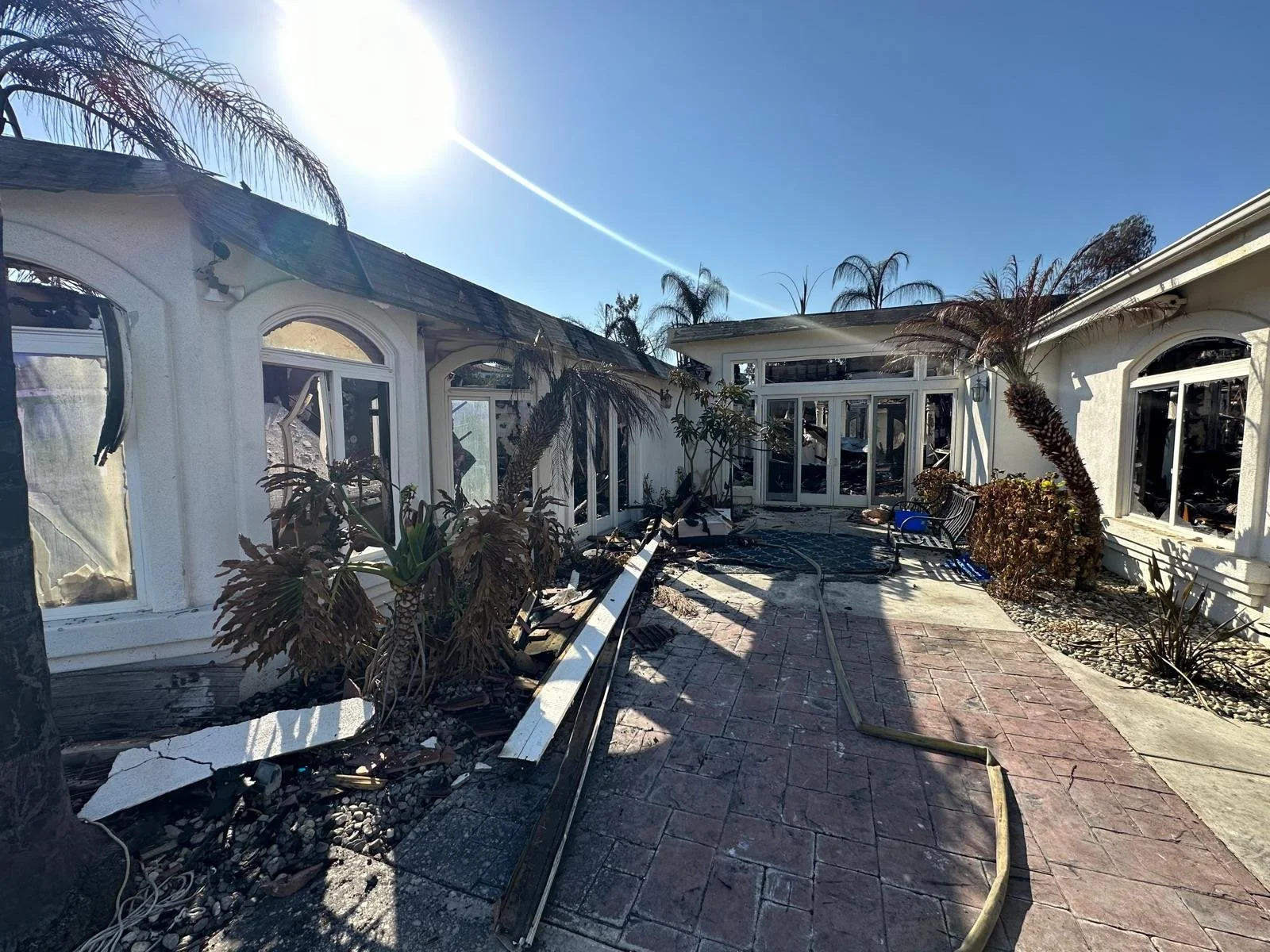 Backyard of a house with extensive fire damage, charred and broken palm trees, broken windows, and debris Strewn across the patio under a clear blue sky.