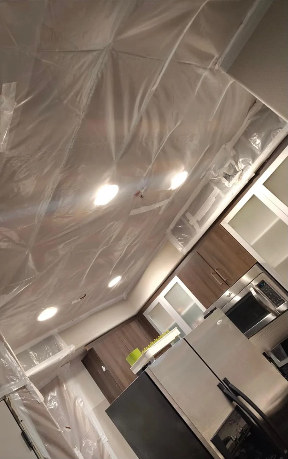 A kitchen under renovation with unpainted drywall and plastic coverings on the ceiling and walls, modern cabinetry, and stainless steel appliances.