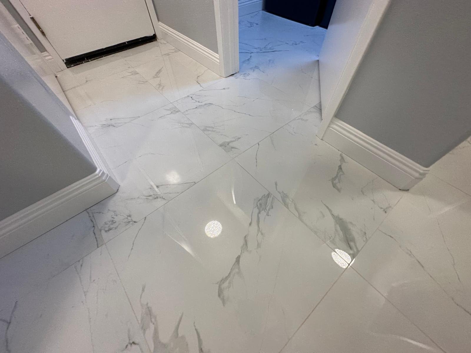 Close-up of a shiny marble floor with grey and white veining, showing reflections of ceiling lights in an indoor setting.