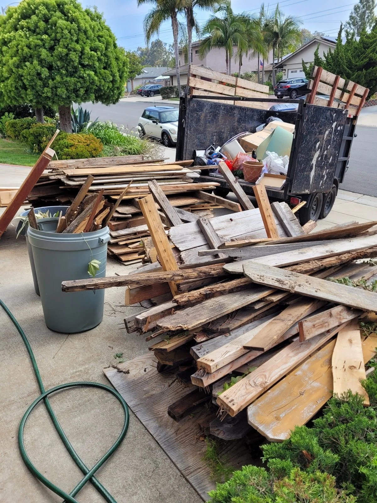 A pile of wooden planks, some stacked on a pallet, with a trash bin nearby filled with more wood and debris. A black trash trailer is filled with assorted trash and debris, parked on a driveway in a suburban neighborhood with trees, houses, and parke