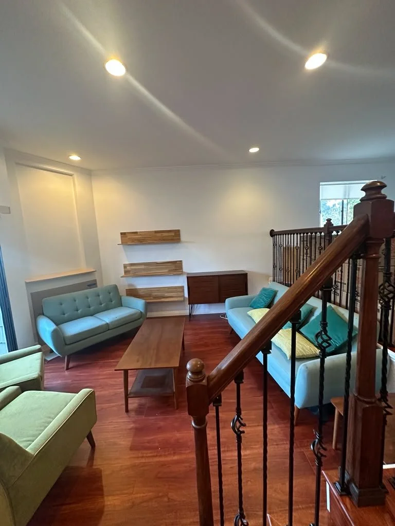 Living room with hardwood floors, light blue and green sofas, wooden coffee table, wooden wall shelves, brown sideboard, staircase with wooden handrail and black metal balusters, window with outdoor view.