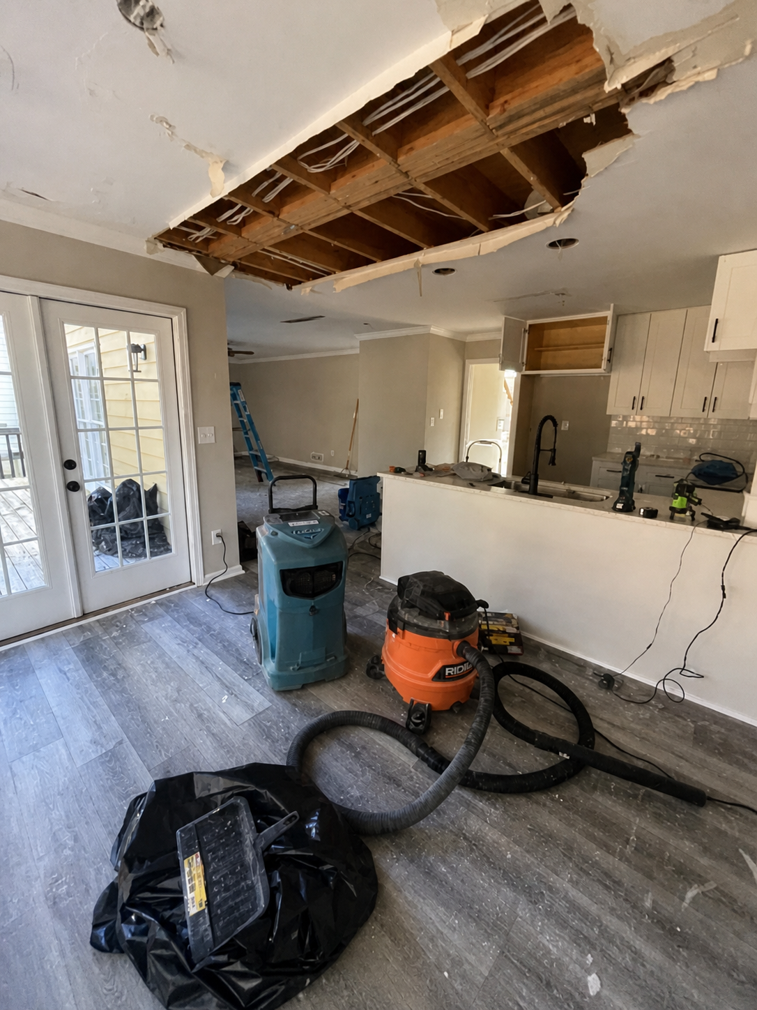 Interior of a house under renovation with a large hole in the ceiling exposing wooden beams and electrical wiring, vacuum and dehumidifier on the floor, and construction tools and materials scattered around.