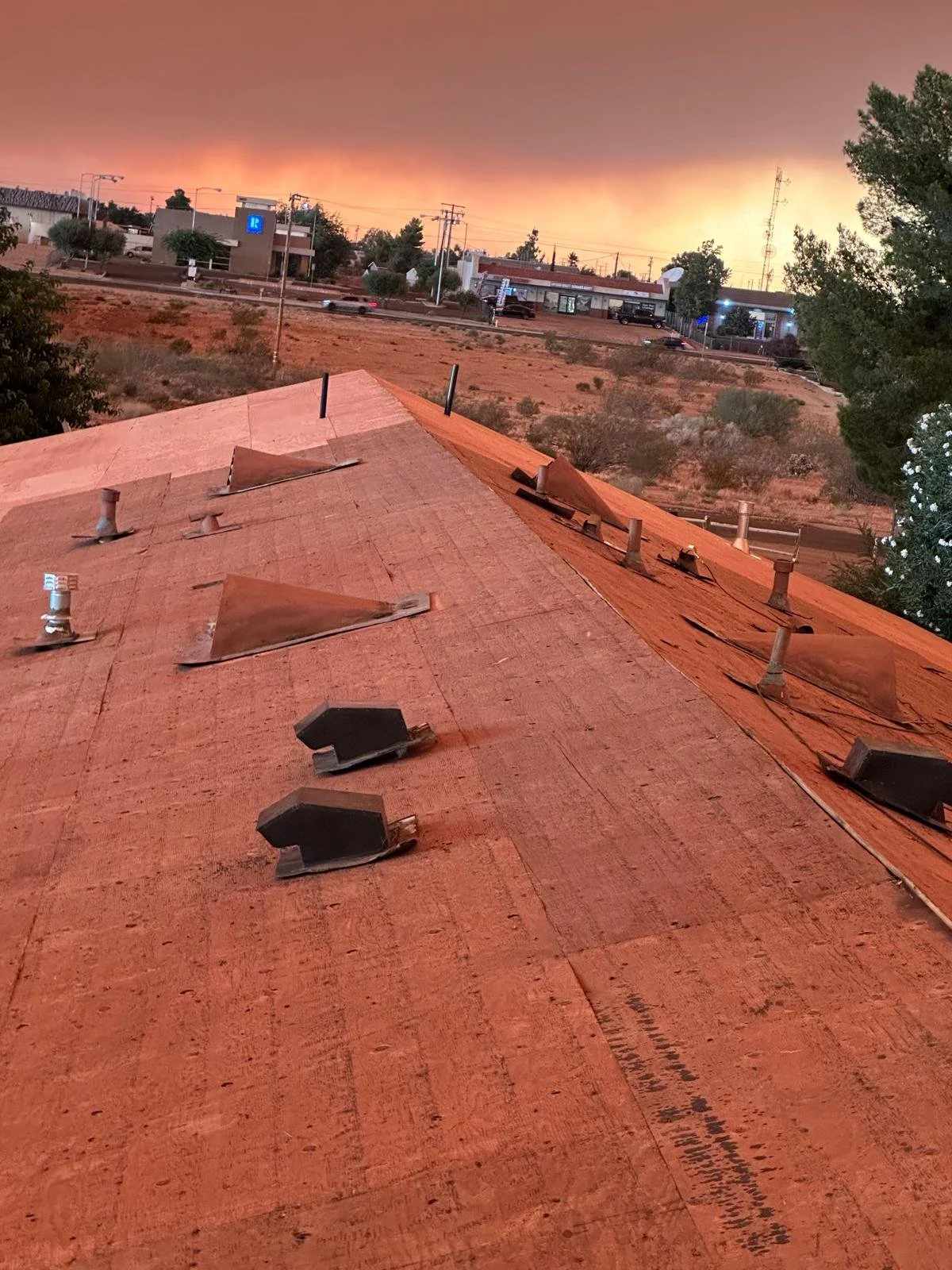 View of a rooftop with shingles and vents in the foreground, with a sunset sky and a street with buildings and trees in the background.