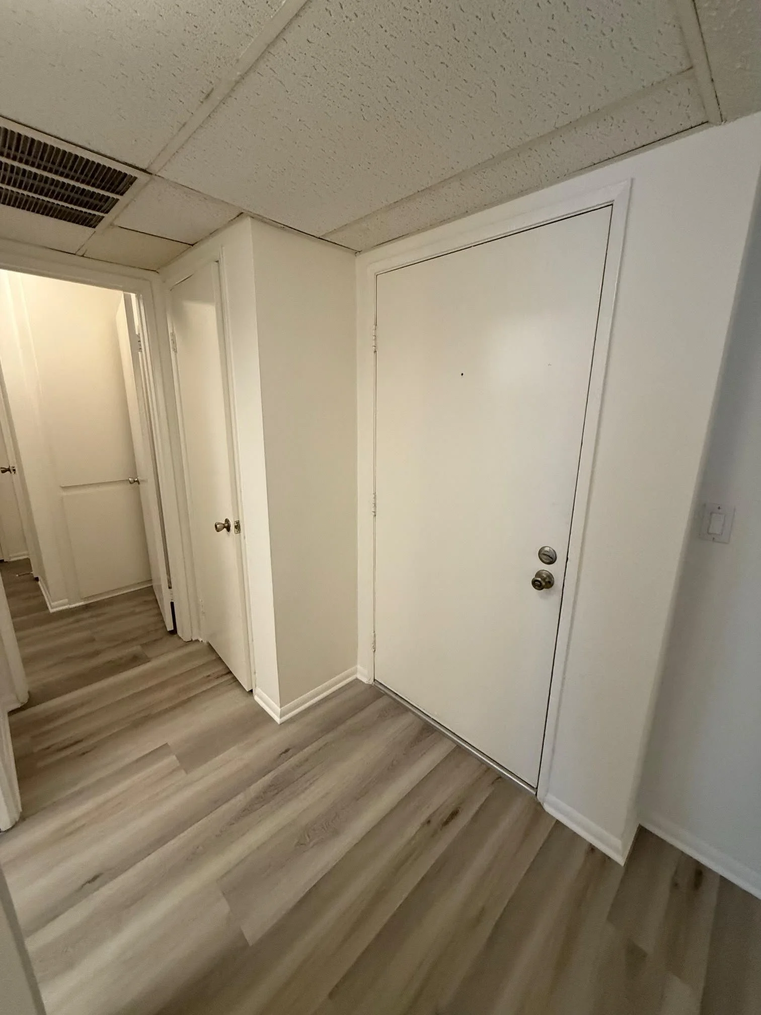 An interior hallway with wooden flooring, white walls, and a white door. The ceiling has textured panels and an air vent. There are trim and molding around the door and walls.
