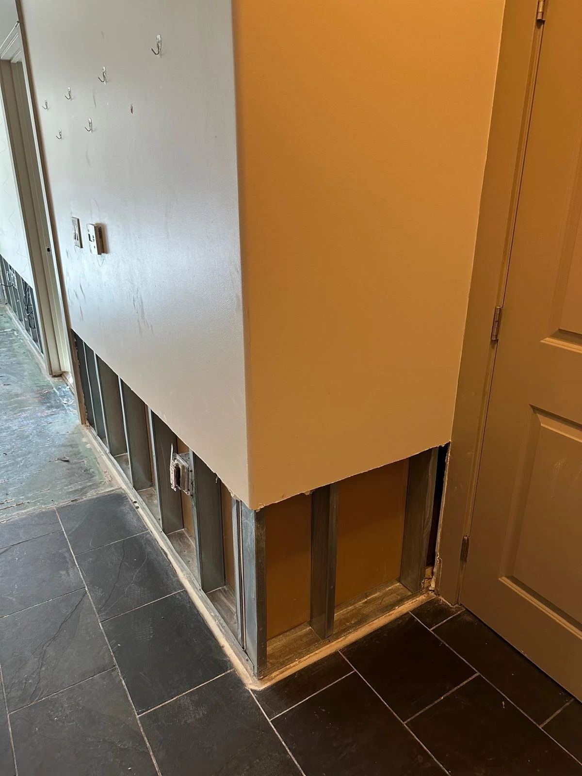 Interior wall undergoing remodeling with exposed wall studs and partially painted drywall, next to a closed beige door, on a black tiled floor.